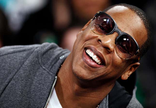 Jay-Z leans to the right, smiling, wearing sunglasses and a hoodie. 