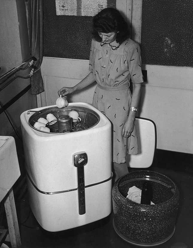 A black and white photo of a woman putting coffee cups into an early dishwasher, with a drum of laundry beside her on the floor.