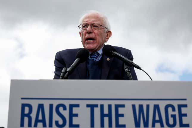 Senator Bernie Sanders speaks at a podium that has a sign reading "raise the wage" in bold, blue font fixed to the front. 