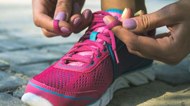 Close-up photo of a person with brown skin and a light purple manicure lacing up bright pink running shoes, which have bright turquoise accents and a white sole.