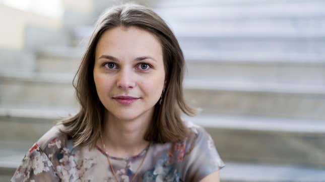 A white woman with shoulder length brown hair and a flowered gray shirt smiles into the camera on a white staircase.