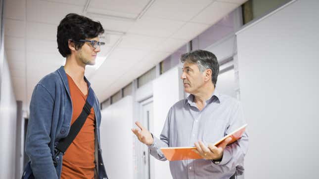 A young man talks with an older man holding a notebook in a hallway