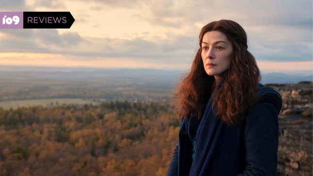 Rosamund Pike's somber Moiraine, wearing a long blue coat and long brown hair. She looks across a vast, gorgeous expanse of medieval landscape.