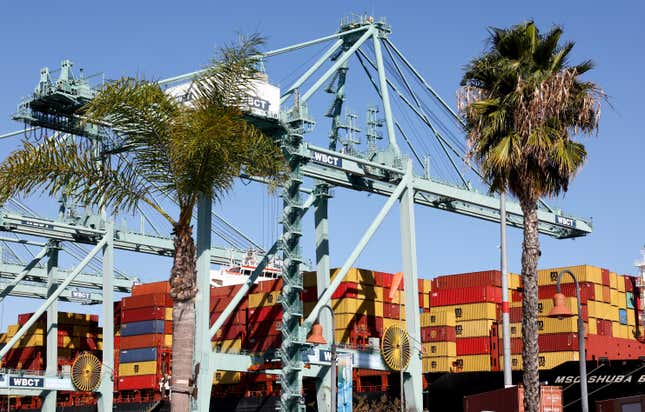 Shipping containers rest on a container ship behind palm trees. 