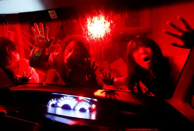 Two actors dressed as zombies press against a car windshield backlit in red.