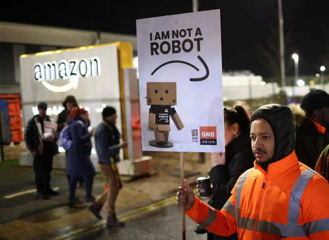 A worker in a bright orange jacket holds a sign reading "I am not a robot" outside of an Amazon warehouse night time. 