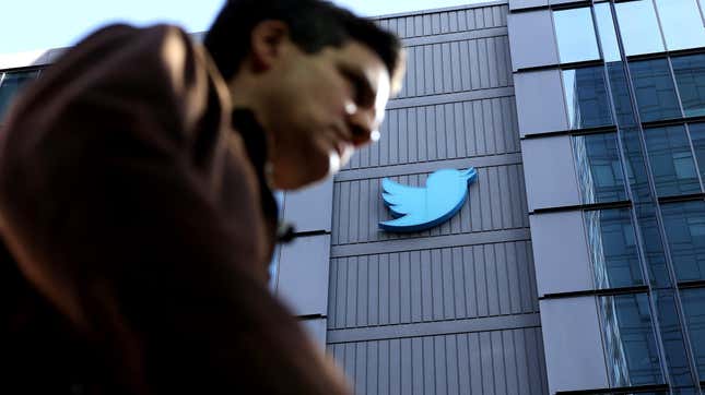 A blurred out man walks past a large building with the blue bird twitter logo.