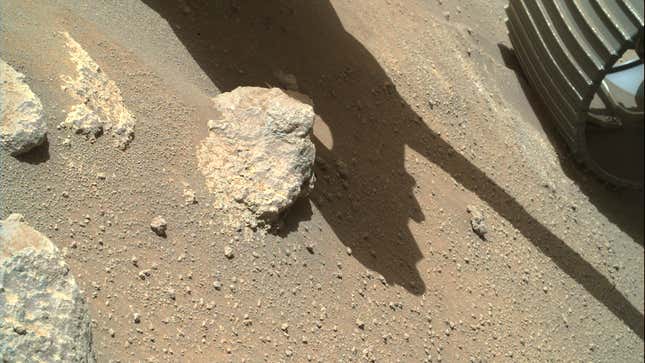 The rust-orange surface of Mars, with a large rock next to the wheel of Perseverance. 