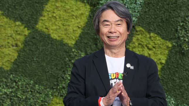 Shigeru Miyamoto smiles while standing in front of a green hedge covered in squares. 