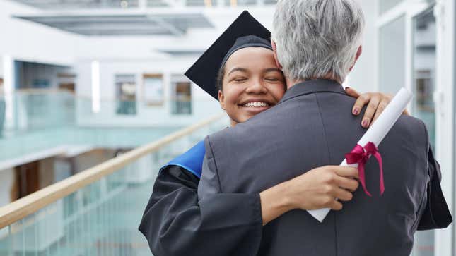 An African American woman wearing a black cap and gown and holding a diploma is seen hugging a grey-haired man wearing a suit, seen from the back