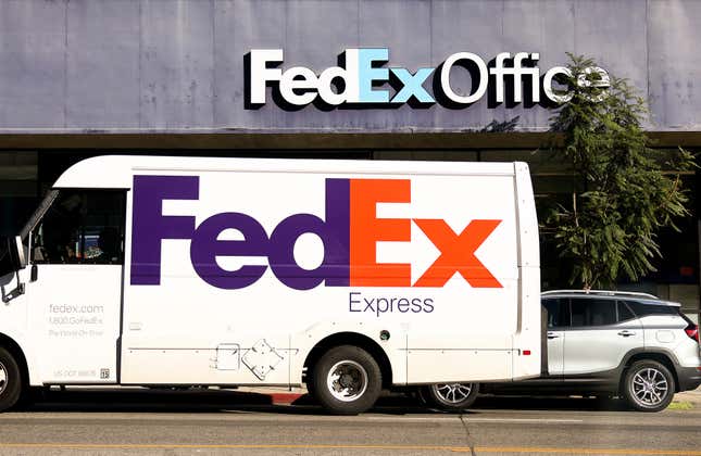 A white FedEx van is parked outside a FedEx Office on a sunny street.
