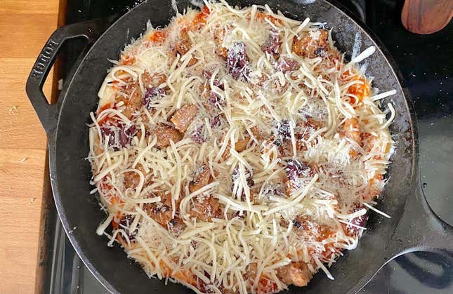 Pizza dough placed on top of the parmesan layer in the cast iron skillet. The pizza is topped with sausage chunk, sliced kalamata olives, shredded parmesan and shredded mozzerella.