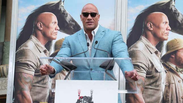 Dwayne Johnson speaking at a lectern in front of two posters for the film "Jumanji"