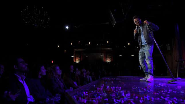 Chris Rock standing on a stage in Brooklyn surrounded by a laughing audience.
