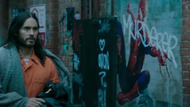 Morbius, in a orange jumpsuit and gray robe, walks past a graffitied picture of Spider-Man in an alley.