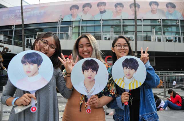 Three fans stand next to each other, smiling and throwing up peace signs while holding three round fans with printed portraits of BTS members Jimin, V, and Suga. Behind them is a stadium with a large poster of BTS.