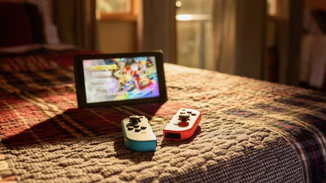 A Nintendo Switch and two detached Joy-Con controllers site on a bed as light comes in from a nearby window.