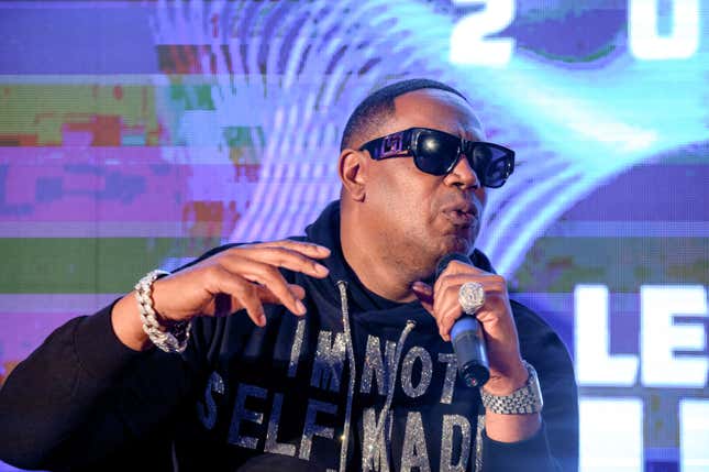 A Black man wearing sunglasses, a black hoodie with sparkly words, and a large watch and diamond ring speaks into a microphone.