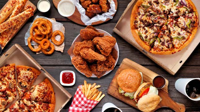 A bunch of fast food—fries, pizza, fried chicken, burgers, onion rings, chicken wings—laid out on a table