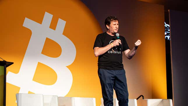 Alex Mashinsky with microphone in hand gestures in front of a large Bitcoin symbol.