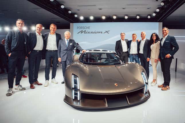 Eight men and one woman, all in business suits, stand around the Porsche Mission X. Overhead rear shot of the Porsche Mission X. Car sits on a white mirrored stage surrounded my stage lights in the rafters A huge digital display behind the silver-gold car reads Porsche Mission X