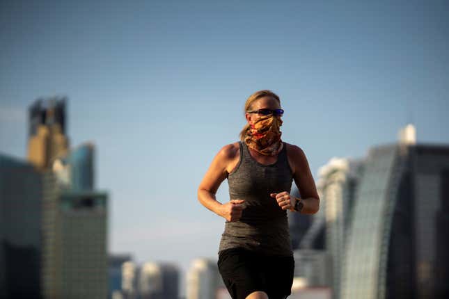 A woman running in black shorts and a grey shirt with an orange buff pulled over her nose and mouth.