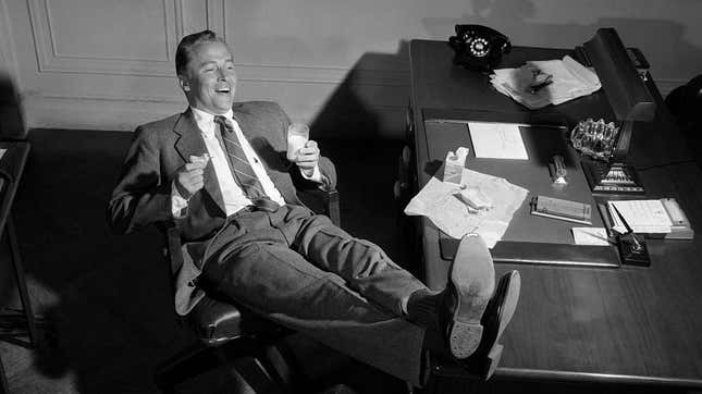 Black and white photo of man laughing with feet up on desk, drinking glass of milk