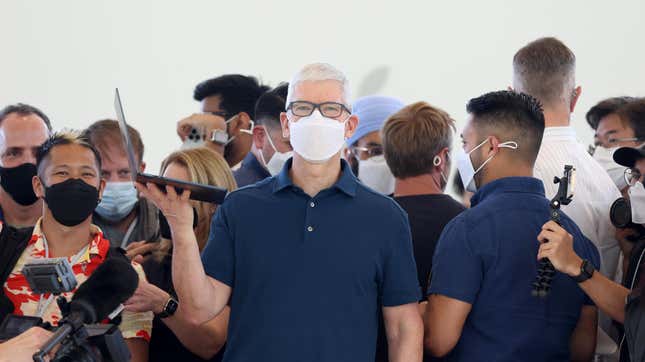 Apple CEO Tim Cook stands in the middle of a throng of people wearing masks, holding a laptop in one hand and staring at the camera.