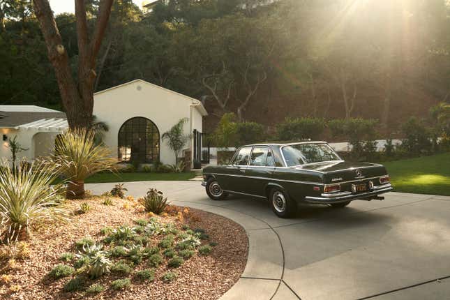 A green 1970 Mercedes is parked on a curved driveway in front of a white house