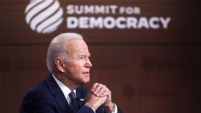 President Joe Biden is sitting clasping his hands. A sign says Summit for Democracy in the background.