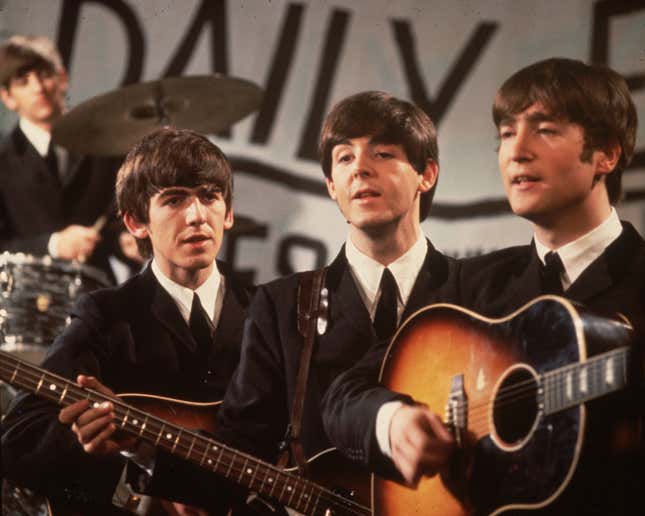 An old photo of the Beatles on stage performing. In the background, blurry, is Ringo Starr. In the foreground, George Harrison to the left, Paul McCartney center, and John Lennon right, playing guitars/bass and singing.