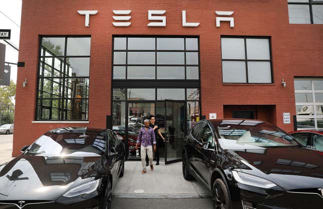 A Tesla service center and show room. The building is red brick with large black windows. Two people are walking out of the building, which is flanked by two black Tesla models.