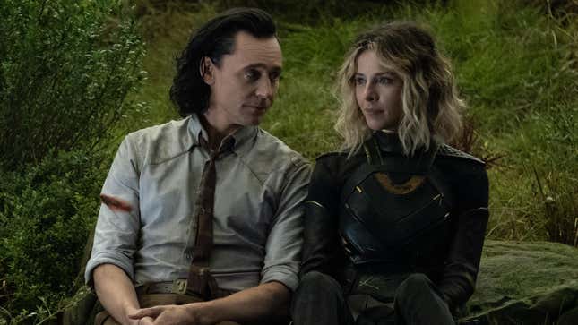 Tom Hiddleston's Loki wears a dirty shirt and tie while sitting next to Sophia Di Martino's Sylvie in her supersuit on the ground.