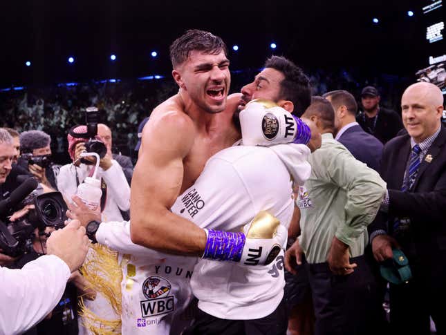 Tommy Fury jumps into the arms of his coach yelling in celebration surrounded by ringside photographers.