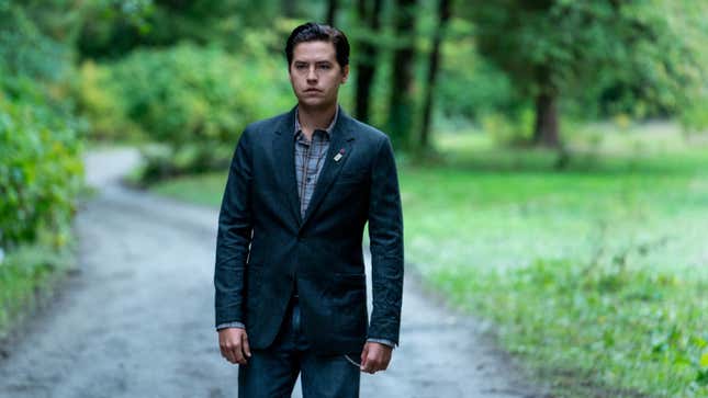 Riverdale's Jughead, dressed in a sharp suit and with slicked back hair, stands alone on a gravel road with trees in the background.