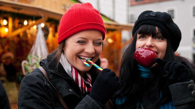 Two women at a Christmas market eating apple and candy in front of a booth.