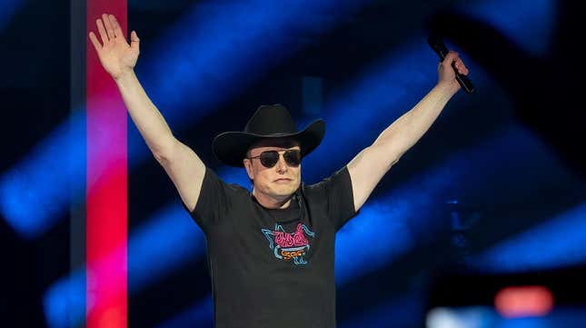 Elon Musk lifts both hands in the air wearing black sunglasses and a black cowboy hat.