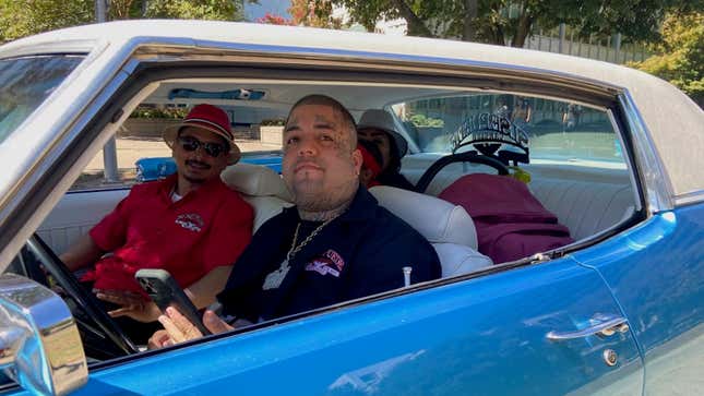 Two men, one heavily tattooed are in the front seat of a blue lowrider car. There is a woman and baby seat in the back.