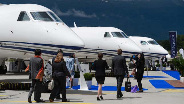 A group of six people walking in front of a row of private jets on the tarmac.