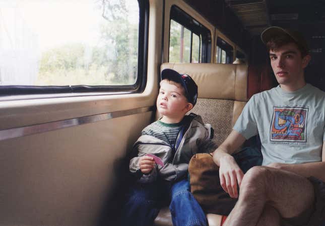An older Nickerson sits next to his younger self on a train looking surprised.