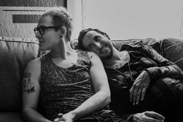 Two men sit on a couch, looking to the left. The man on the right, Trystan, holds his pregnant belly.