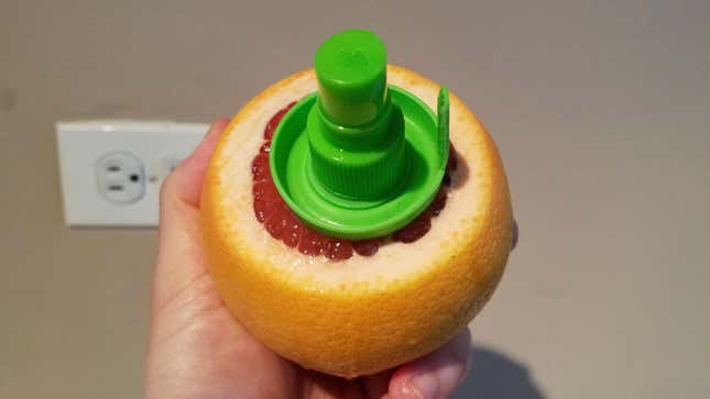 hand holding up a grapefruit with the end cut off and the citrus sprayer screwed in, with outlet on wall visible in background