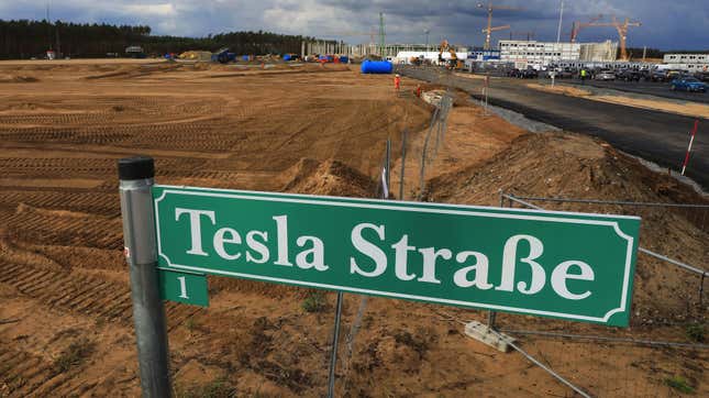 A green street sign reading "Tesla Street" stands in the foreground while a desolate, dirt field stretches behind it. A factory is under construction in the distance on the right, and a stand of tress sits far off on the left. The sky is cloudy and looks as if it's about to rain.