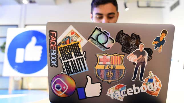 a man on a laptop with several stickers with Facebook and Instagram logos on it sits next to a giant sign with the Facebook thumb up