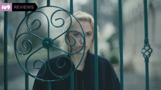 A woman with light blonde hair, wearing a black jacket, stares through an ornate fence in a scene from Watcher.
