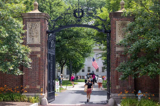 People walk through the wrought iron and brick gate on Harvard Yard at the Harvard University campus in summer.
