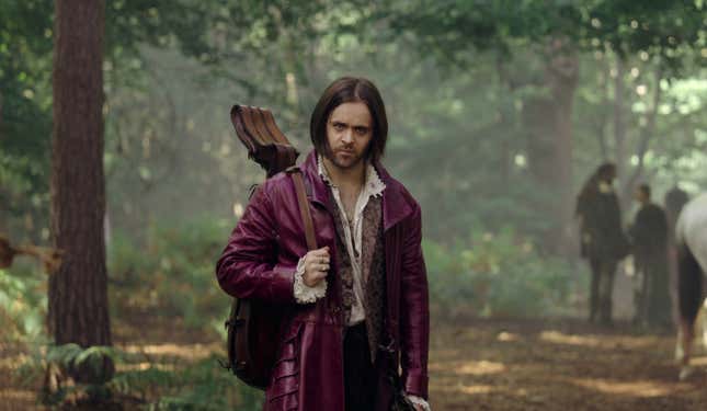 Joey Batey as Jaskier wearing a purple leather coat and carrying a lute in The Witcher season three. 