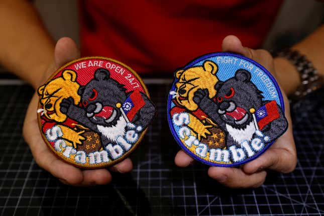 A person's hands, holding two versions of the Taiwanese patches, both showing a black bear with red eyes punching Winnie the Pooh. One says We are open 24/7 and the other says Fight for freedom. Both say Scramble!