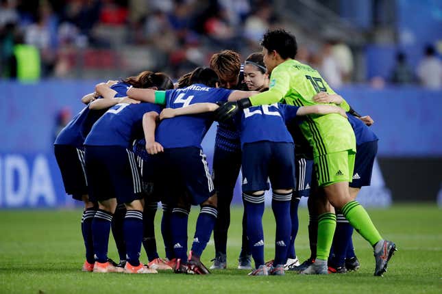 A group of Asian women in matching royal blue shirts and black shorts with blue socks huddle at centerfield on a soccer pitch.