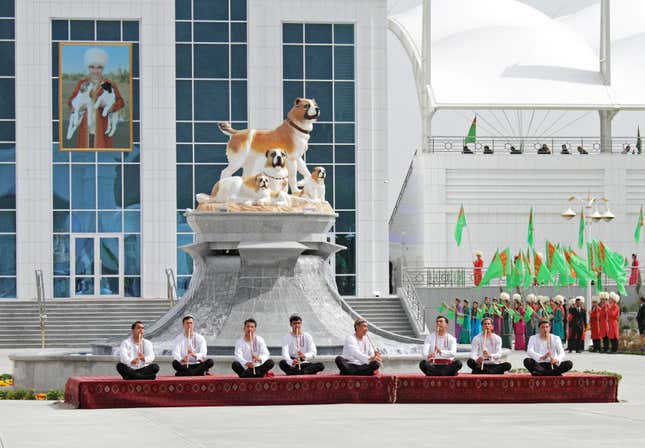 A statue of a family of dogs rises on a white plinth. In front is a line of musicians playing a flute-like instrument. In the background is a modern-looking building with a group of individuals holding Turkmenistan flags.
